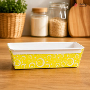 Rectangular baking dish with yellow and white pattern on a wooden surface, with jars and a plant in the background.