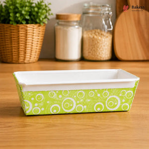 Rectangular baking dish with green and white pattern on a wooden surface, with jars and a plant in the background.