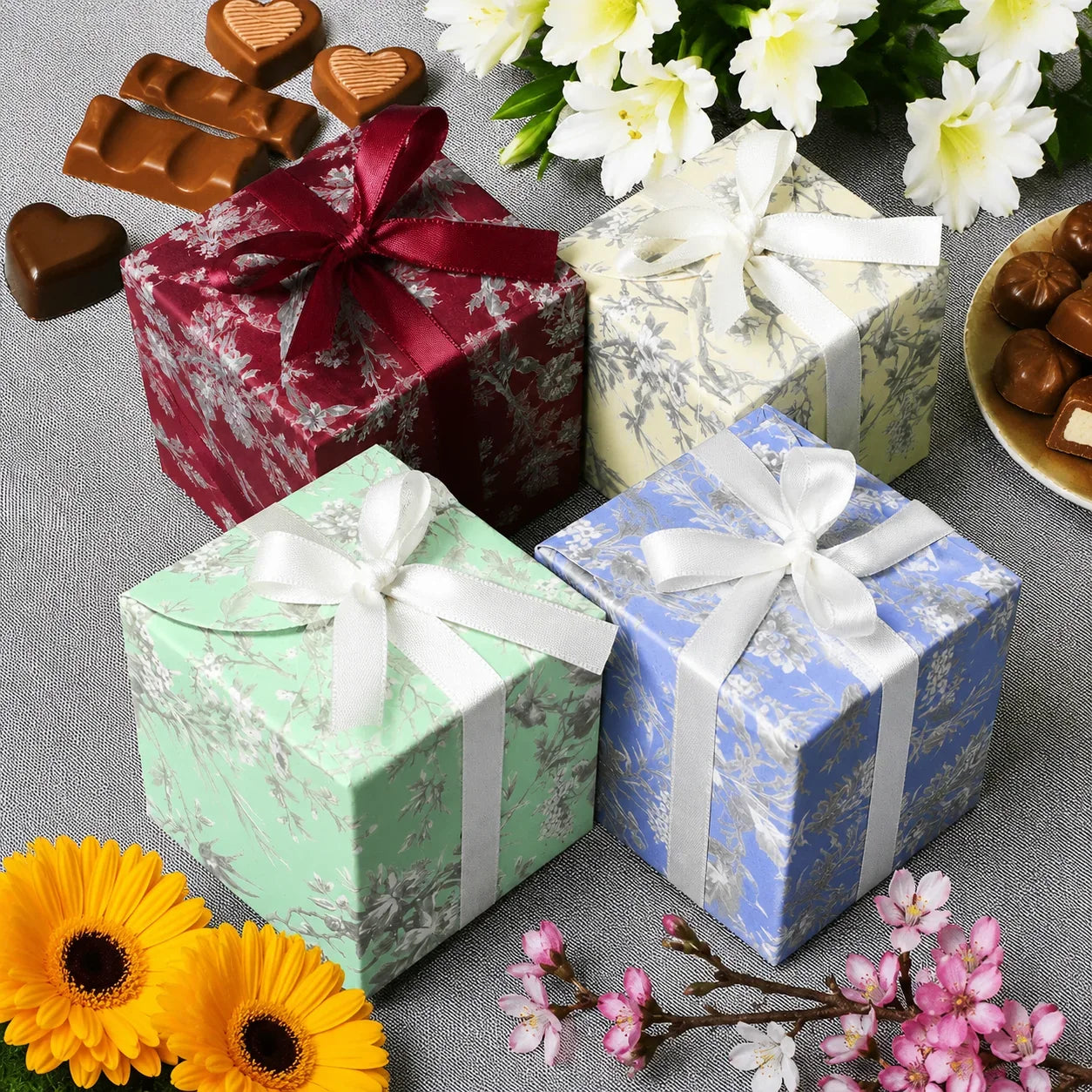 Four small gift boxes with ribbons on a gray surface with flowers and chocolates.
