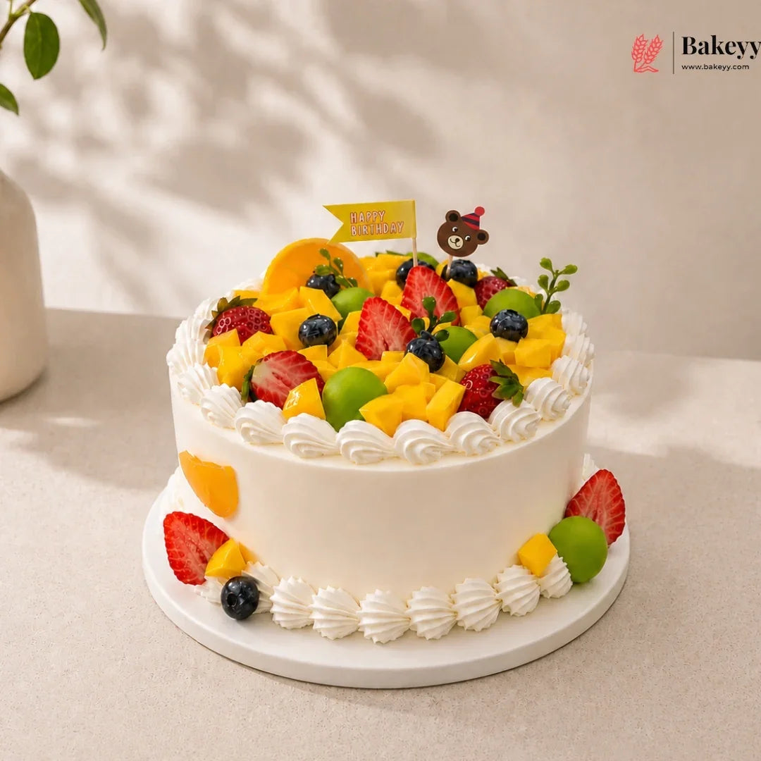 Fruit cake with white frosting and colorful fruits on a light background, featuring the brand 'Bakeyy'.