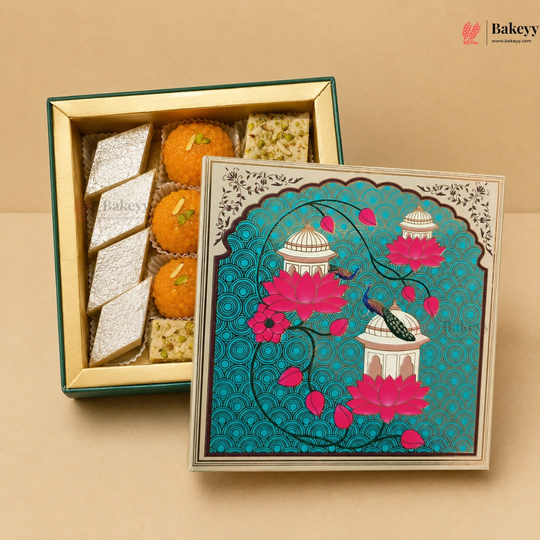 An open decorative gift box of Indian sweets and snacks, with one side showing laddooes and pistachio-filled squares, and the lid leaning in front featuring a turquoise background with pink lotus flowers, domes, and a peacock. The box is photographed on a beige studio background.