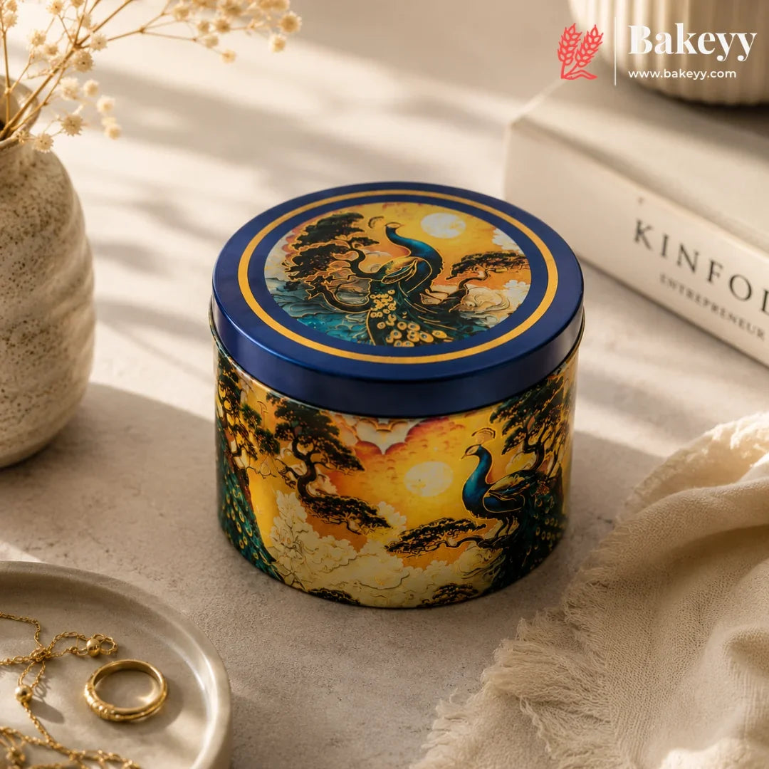A decorative round metal tin with a blue lid and a colorful peacock landscape illustration, styled on a beige surface with a book, jewelry, and dried flowers in the background.