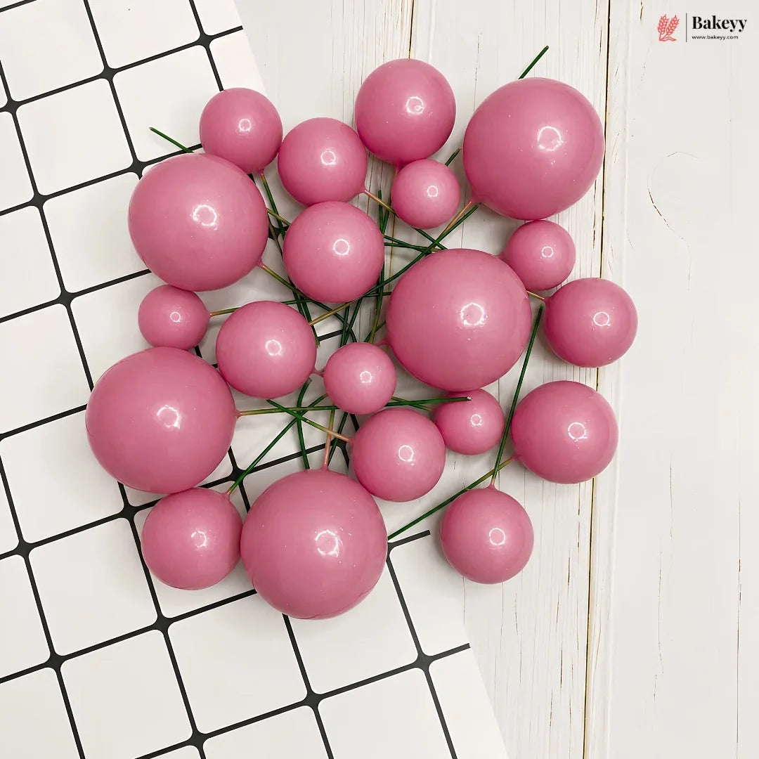 A cluster of glossy pink decorative beads or pom-poms on thin green stems, arranged on a white surface with a black grid pattern and a white wood plank background.