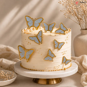 Decorative cake with butterfly toppers on a marble stand, with a vase and textured fabric in the background.