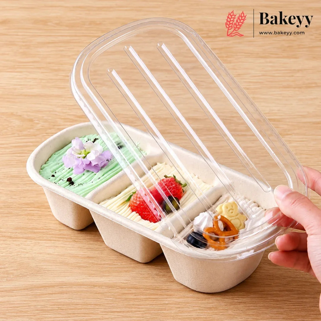 A three-compartment paperboard dessert tray filled with colorful cakes or pastries, shown on a wooden table with a hand lifting a clear plastic lid. The Bakeyy logo appears in the top right corner.