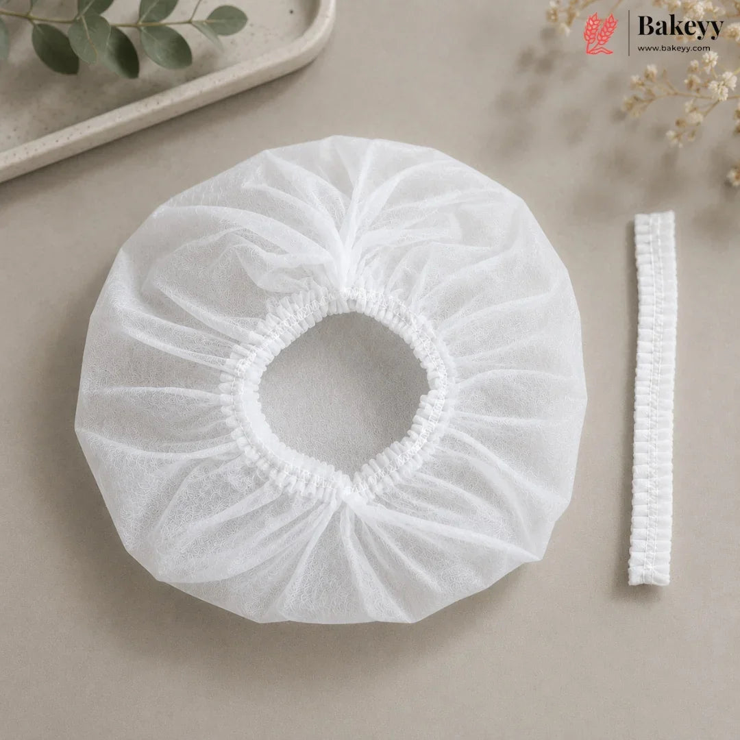 A white disposable bakery dust cover with an elasticized center and a matching elastic band, displayed on a neutral beige surface with decorative greenery.