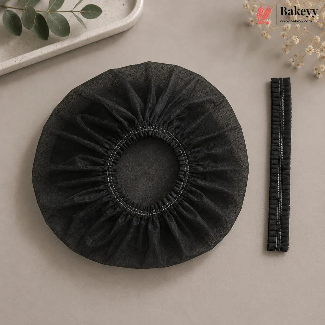 A black stretch fabric hair cap or bonnet with a gathered elastic center, shown on a beige surface with a matching black zipper strip beside it. Decorative greenery and flowers appear in the corners, and the Bakeyy logo is visible in the top right.