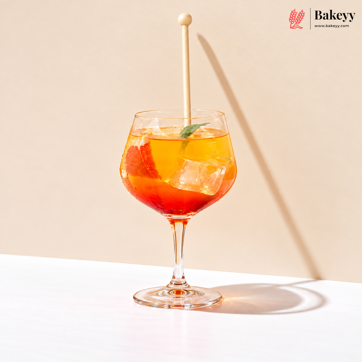 A clear stemmed glass filled with an orange-red drink, ice cubes, a green leaf garnish, and a wooden spoon, set on a white surface against a beige background with a strong shadow. The Bakeyy logo appears in the top right.