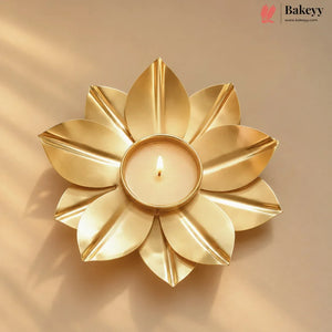 A gold-toned flower-shaped candle holder with a lit tealight candle in the center, set against a warm beige background with soft shadows. The Bakeyy logo is visible in the top right corner.