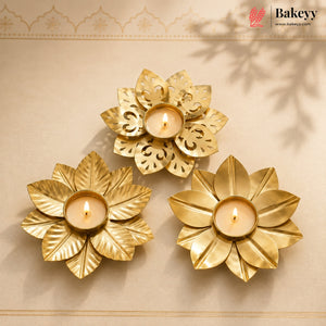Three gold-toned flower-shaped candle holders with lit tealight candles, arranged on a beige surface with a decorative border and soft leaf shadows.