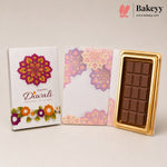 A Diwali-themed chocolate gift set from Bakeyy, featuring a white box with a magenta mandala and floral design, an open matching envelope, and a gold-trimmed chocolate bar.