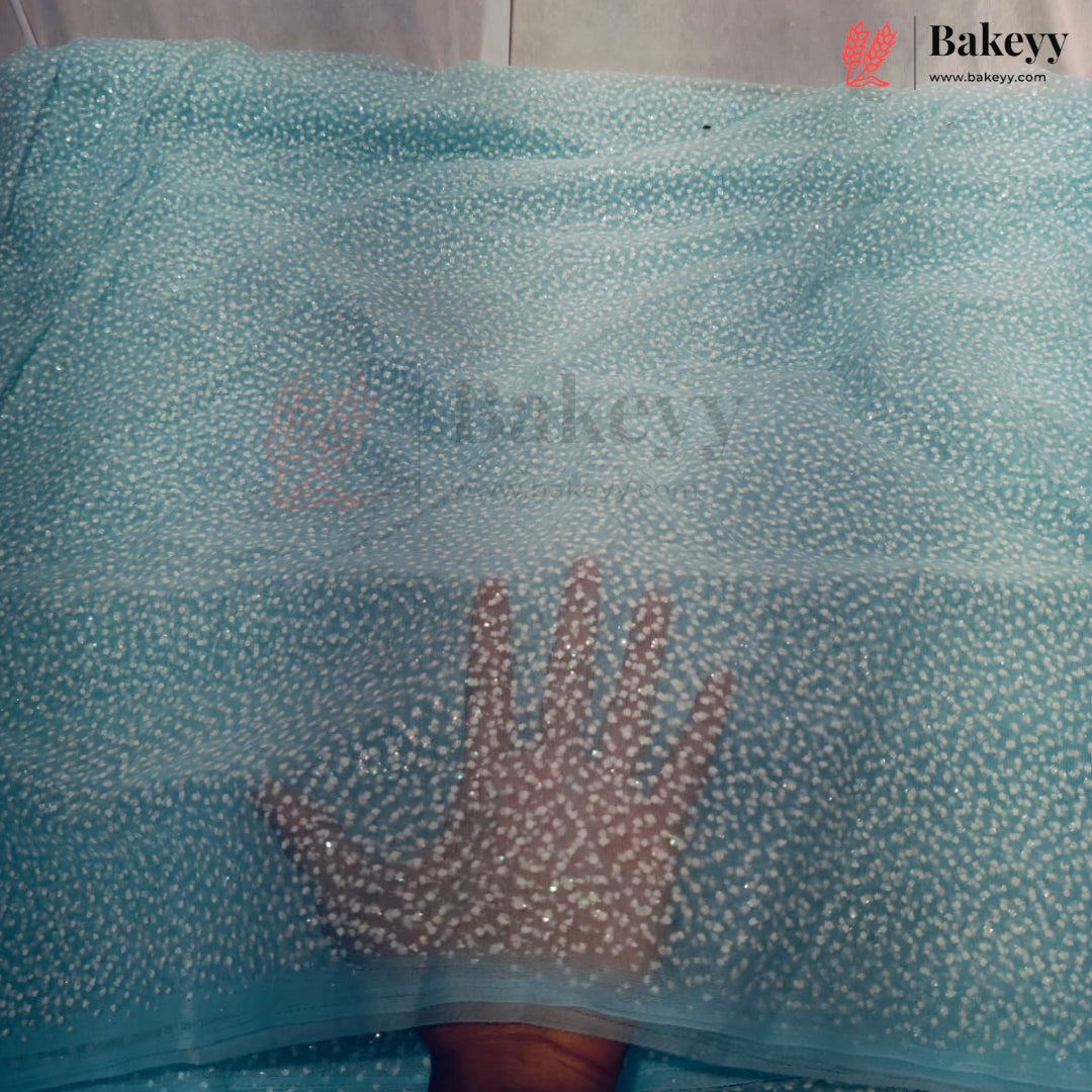 A close-up of a light blue sheer fabric heavily embellished with small white sequins, with a hand placed underneath to show transparency and texture. A watermark for 'Bakeyy' is visible on the fabric and in the top right corner.