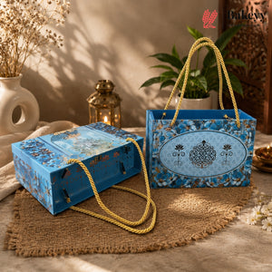 Two decorative blue bakery gift boxes with rope handles, styled on a burlap mat with dried flowers, a lantern, and a potted plant in a warm indoor setting.