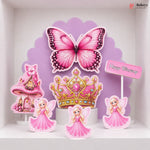 A set of pink fairy-themed birthday cake toppers displayed on a white shelf against a purple scalloped backdrop. The set includes a large pink butterfly, a pink and gold tiara, three fairy figures in pink dresses, a pink castle, and a small 'Happy Birthday' sign.