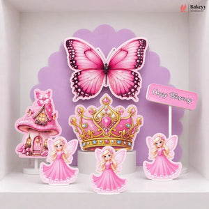A set of pink fairy-themed birthday cake toppers displayed on a white shelf against a purple scalloped backdrop. The set includes a large pink butterfly, a pink and gold tiara, three fairy figures in pink dresses, a pink castle, and a small 'Happy Birthday' sign.
