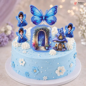 A round light blue frosted birthday cake decorated with a large blue butterfly topper, three fairy figurines, a small fairy house topper, and white piped rosettes, shown on a white cake board against a softly blurred pastel background.