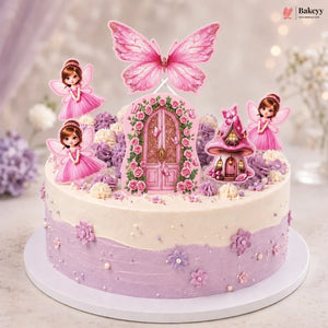 A round fairy-themed birthday cake decorated with pink and lavender frosting, floral details, and figurines including two pink fairy characters, a butterfly topper, a flower door, and a small fairy house. The cake sits on a white round base against a softly blurred pastel background.