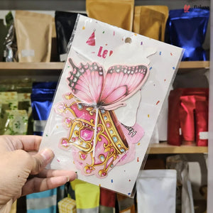 A hand holds a packaged decorative sticker or decal featuring a pink butterfly and ornate gold-and-pink illustration on a white backing card, with shelves of colorful pouches in the background.