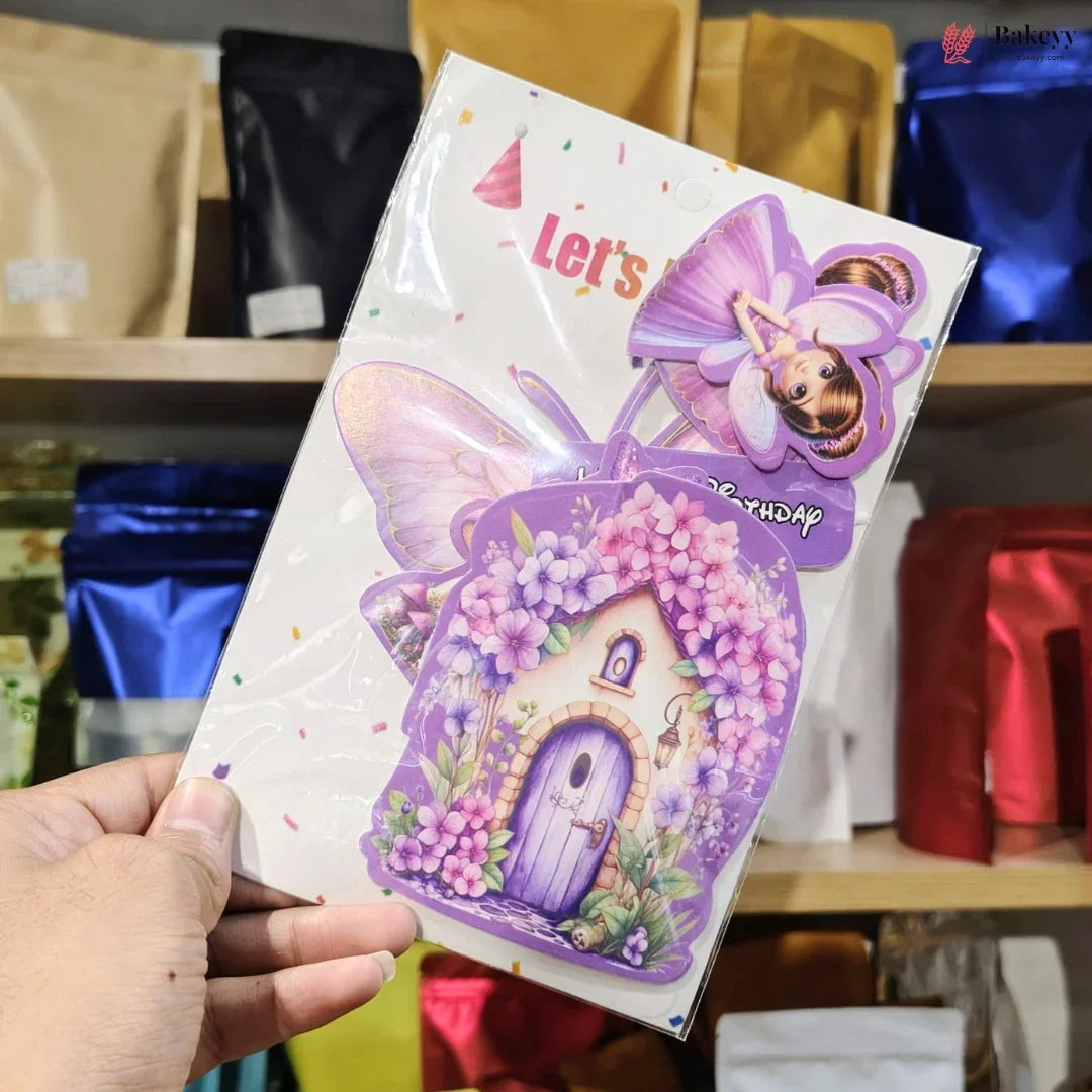 A hand holds a packaged fairy-themed item with a white backing card featuring a purple fairy house, flowers, and a fairy illustration. The background shows shelves with colorful pouches in a retail or storage setting.