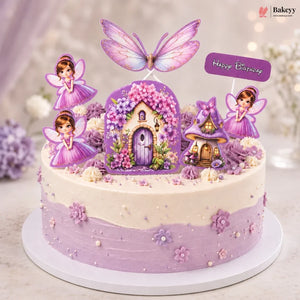 A round birthday cake decorated with lavender and cream frosting, fairy figurines, purple flowers, two small fairy houses, and a butterfly topper. The cake sits on a white round board against a softly blurred indoor background with flowers and warm lighting.