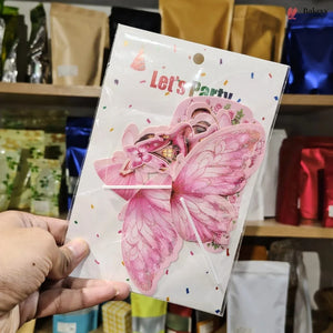 A hand holds a packaged pink butterfly-shaped decorative item with glittery wings and a 'Let's Party' print on the front, photographed in front of store shelves.