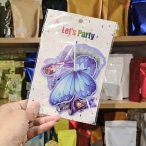 A hand holds a packaged party decoration set featuring a fairy and butterfly design with the text 'Let's Party' on a white backing card, photographed in front of shelves of colorful pouches.