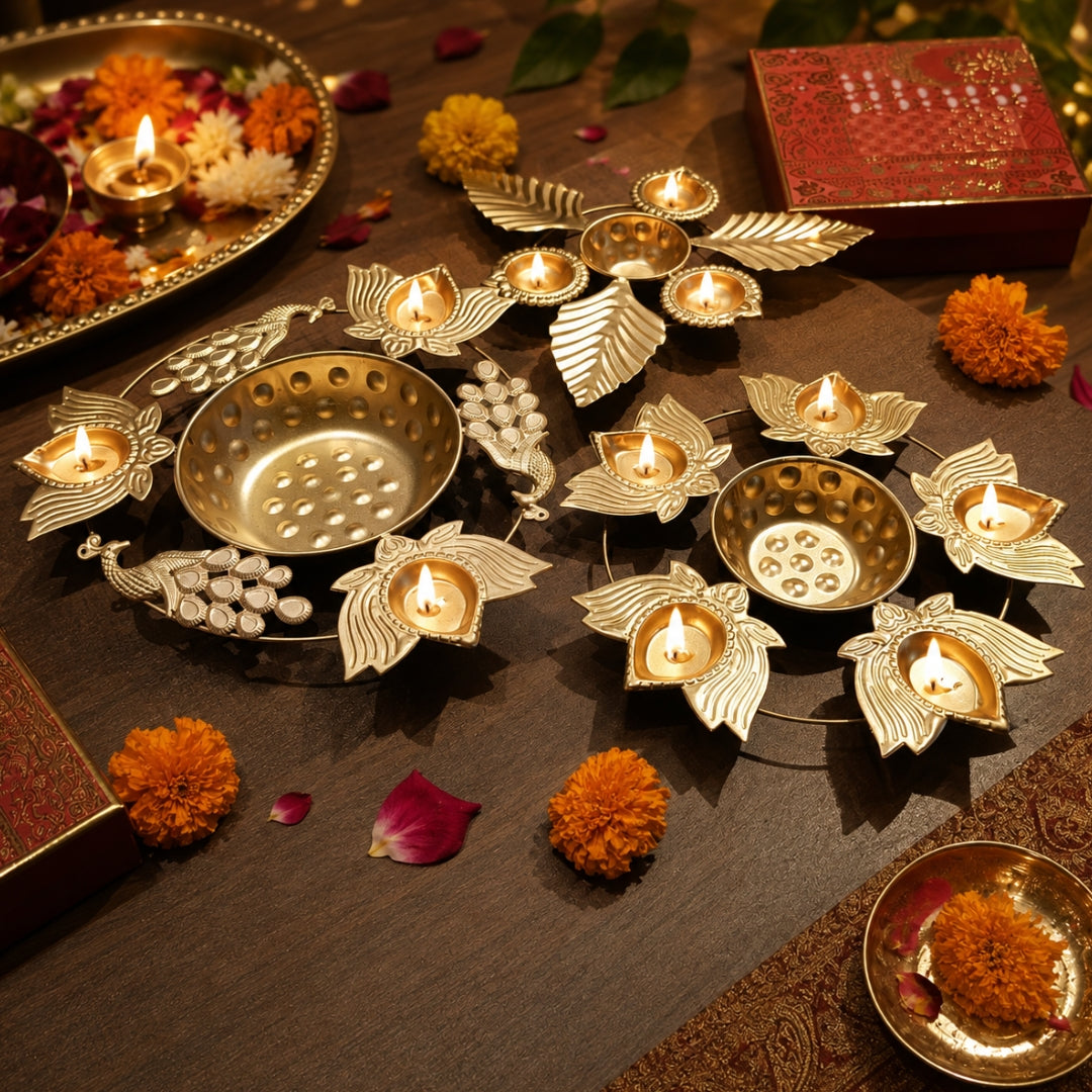 A styled flat-lay of ornate brass and gold-toned diya holders arranged on a dark wooden surface, with lit tealight candles, marigold flowers, and decorative props around them.