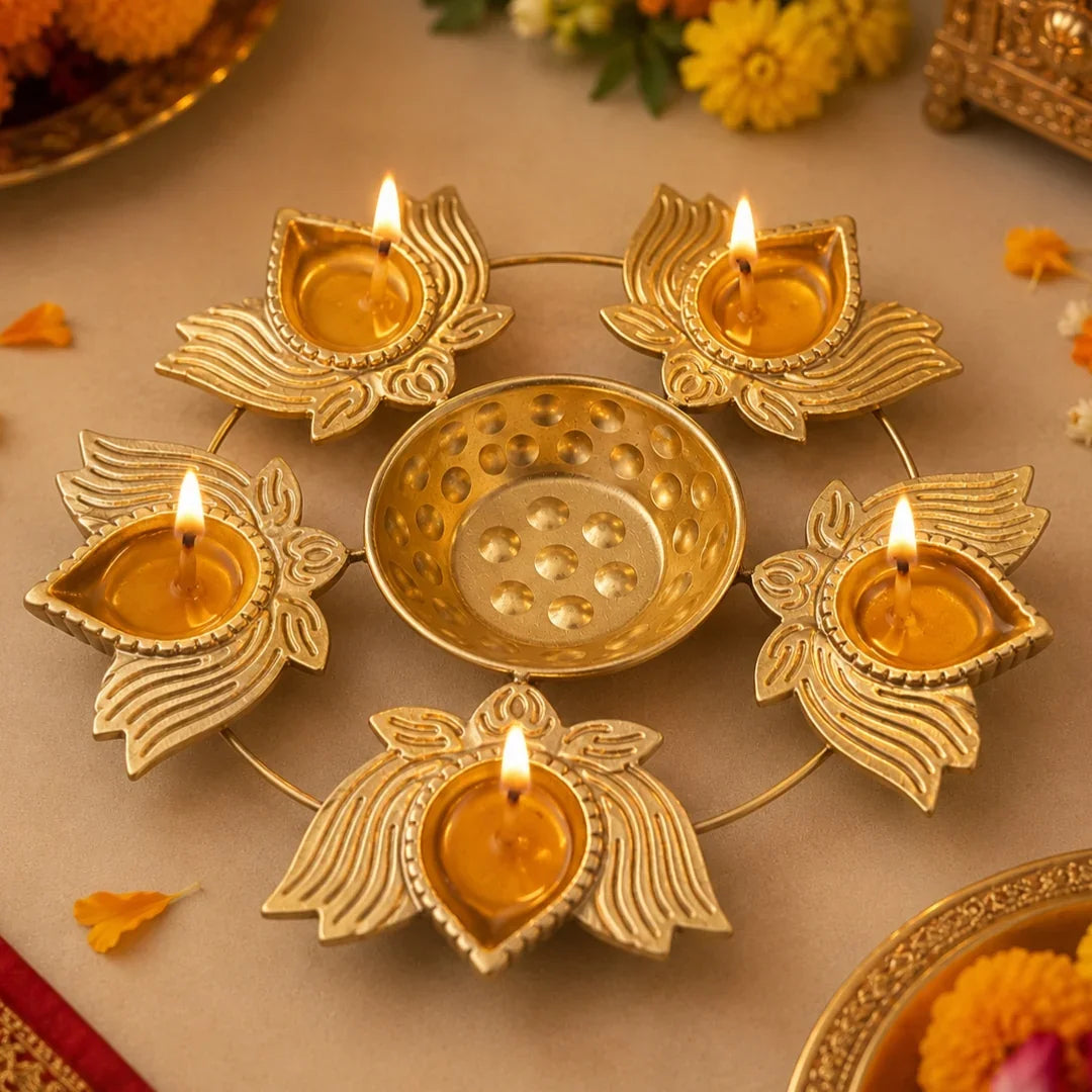 A brass diya light holder with five lotus-shaped candle holders and a central bowl, all lit with small flames, arranged on a beige surface with decorative flowers and petals.