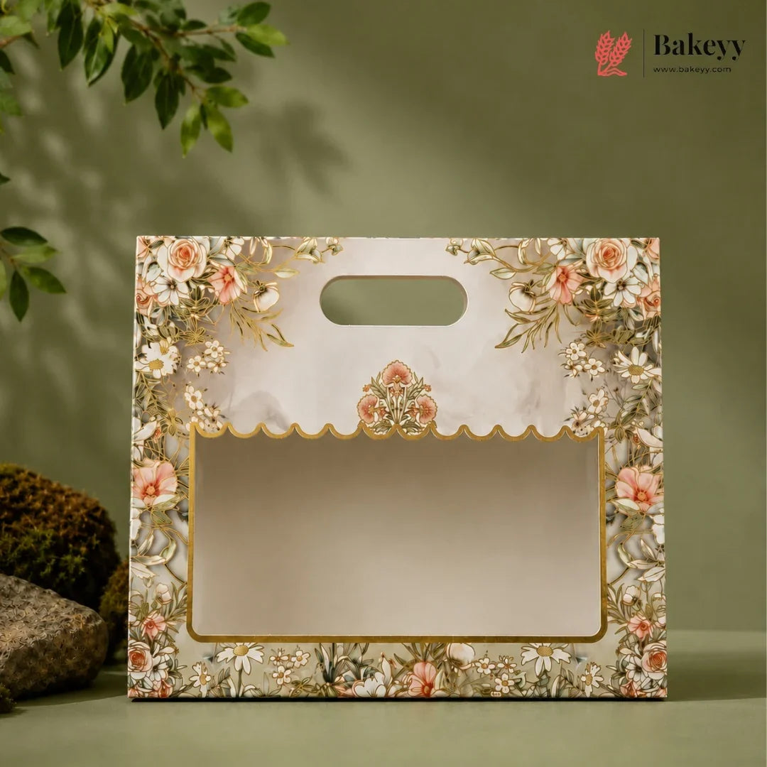 A decorative bakery gift bag with a floral print border, gold trim, and a scalloped window frame, shown on a muted green background with a leafy plant and rocks as props.