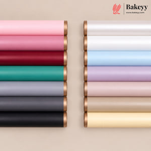 Two vertical stacks of rolled bakery wrap or ribbon in assorted colors, including pink, red, teal, gray, black, white, light blue, lavender, blush, beige, and pale yellow, each with metallic rose-gold ends. The Bakeyy logo and website appear in the top right on a warm beige background.