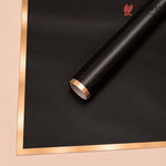 A roll of matte black wrapping paper or vinyl with a metallic rose gold edge, shown diagonally on a beige background with a matching flat sheet of the same material.