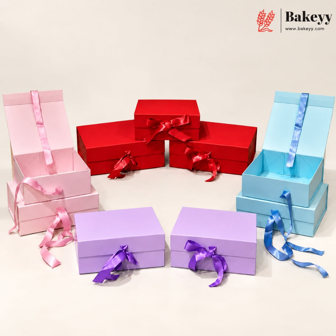 Six colorful bakery gift boxes in pink, red, light blue, and lavender purple, arranged on a light beige background. Some boxes are open, showing interior compartments, and several are tied with satin ribbon bows in matching colors.