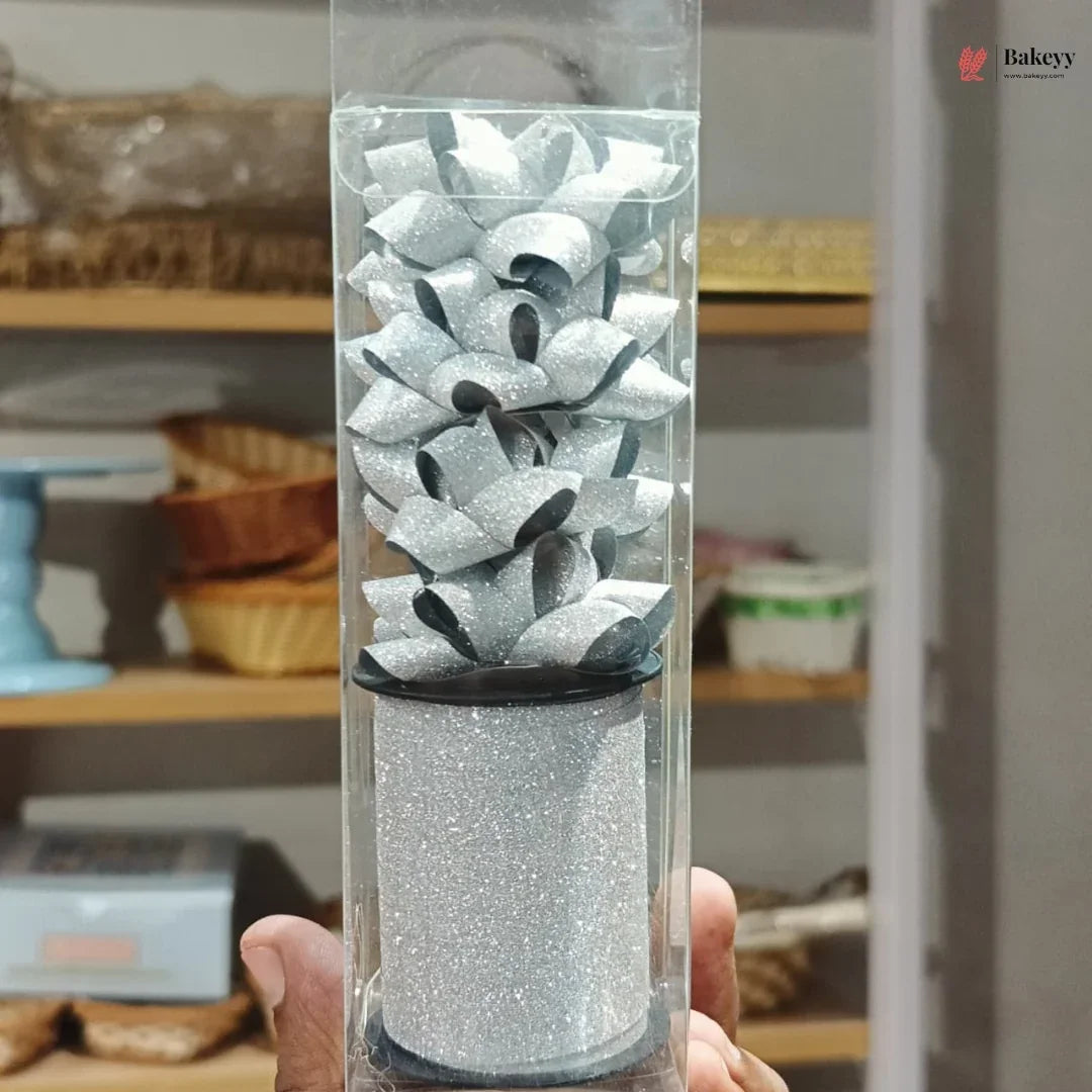 Decorative cake topper with silver and black design in a clear packaging tube, held in front of a kitchen shelf.