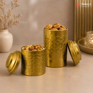Two gold cylindrical metal tins with embossed geometric patterns, one open and one closed, filled with assorted nuts and sweets on a beige tabletop with warm decorative background elements.