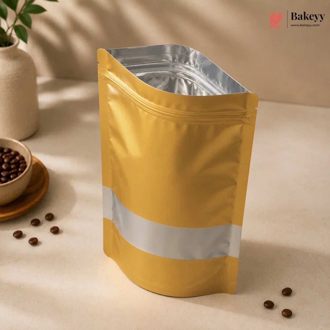 A gold stand-up pouch with a silver reflective band and silver inner lining, standing on a beige surface with coffee beans and a small vase in the background.
