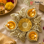 A brass diya holder with five butterfly-shaped candle holders arranged around a central bowl, lit with five burning candles, on a beige fabric background with decorative flowers and a tray in the background. The Bakeyy logo is visible in the top right corner.