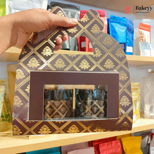 A hand holds a decorative brown and gold patterned gift box with a clear window displaying packaged items inside, set against a shelf background with colorful pouches.