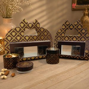 A set of traditional Indian mithai packaging boxes and tins with a dark brown and gold geometric pattern, displayed on a wooden surface with decorative nuts and a warm background.
