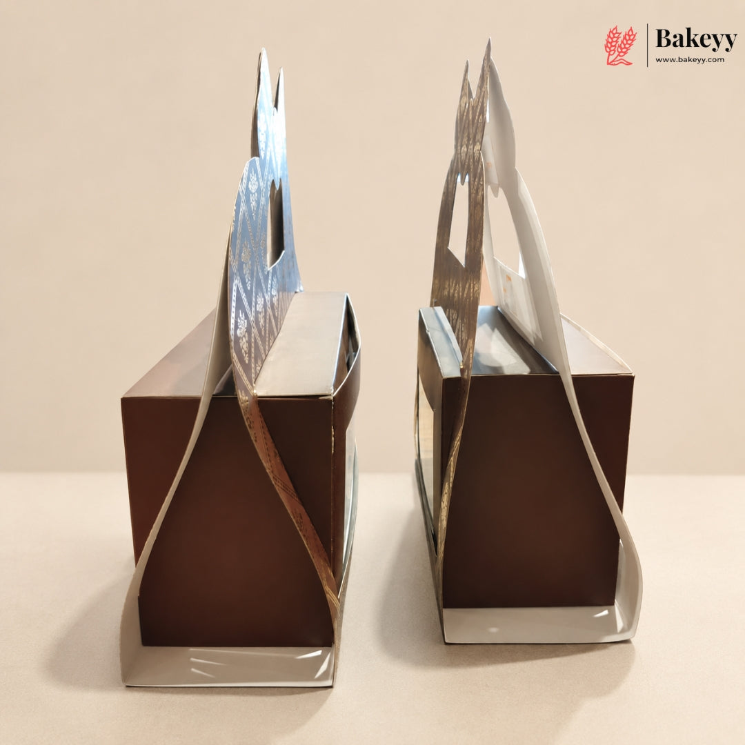 Two square brown bakery boxes with decorative silver and white paper handles, shown on a plain beige background. A Bakeyy logo is visible in the top right corner.