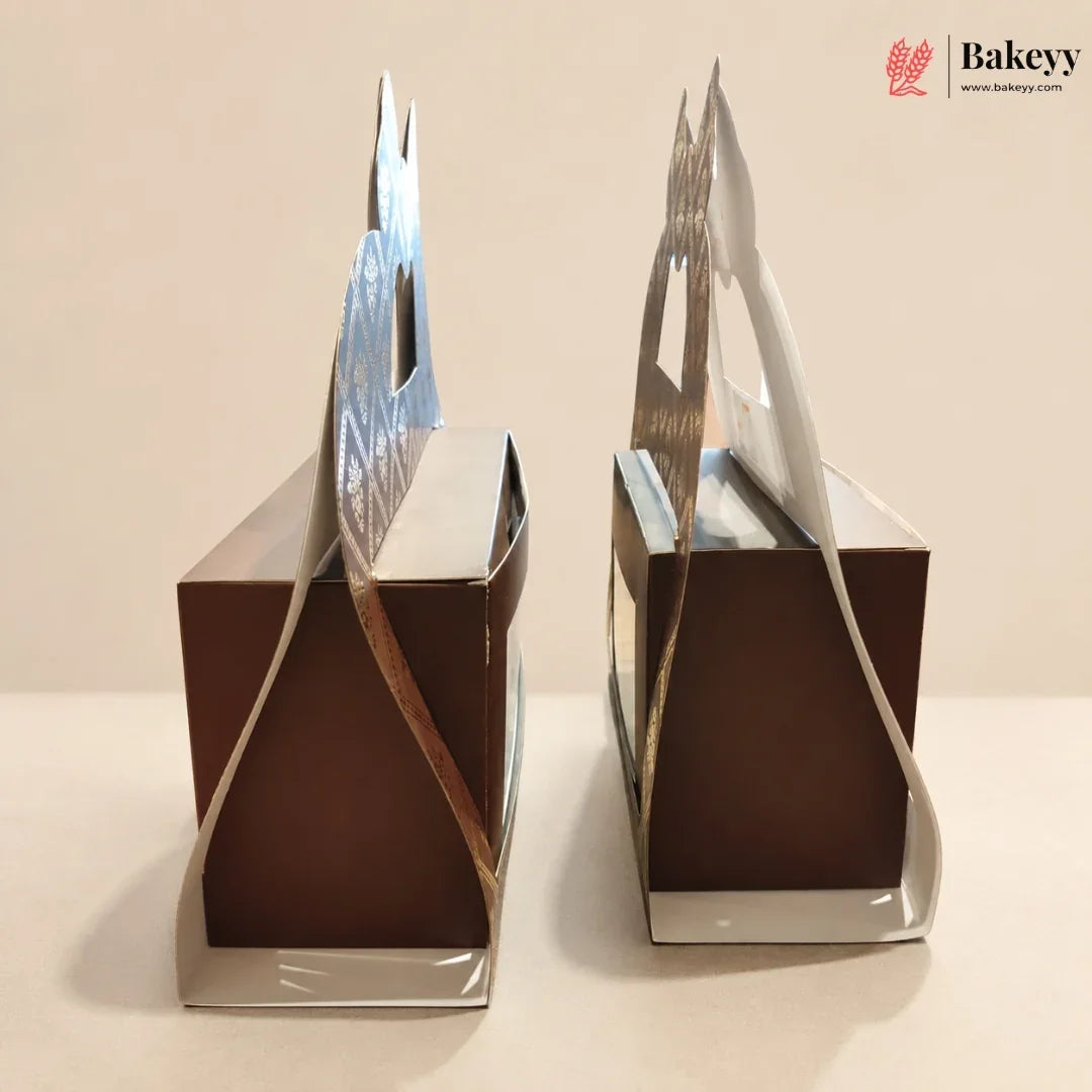 Two square brown bakery boxes with decorative silver and white paper handles, shown on a plain beige background. A Bakeyy logo is visible in the top right corner.