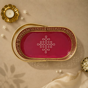 An oval decorative tray with a gold metal frame and handle, featuring a deep magenta-pink interior with a white geometric pattern in the center. The tray is styled on a beige surface with a gold candle holder, small white flowers, and a piece of cream fabric.