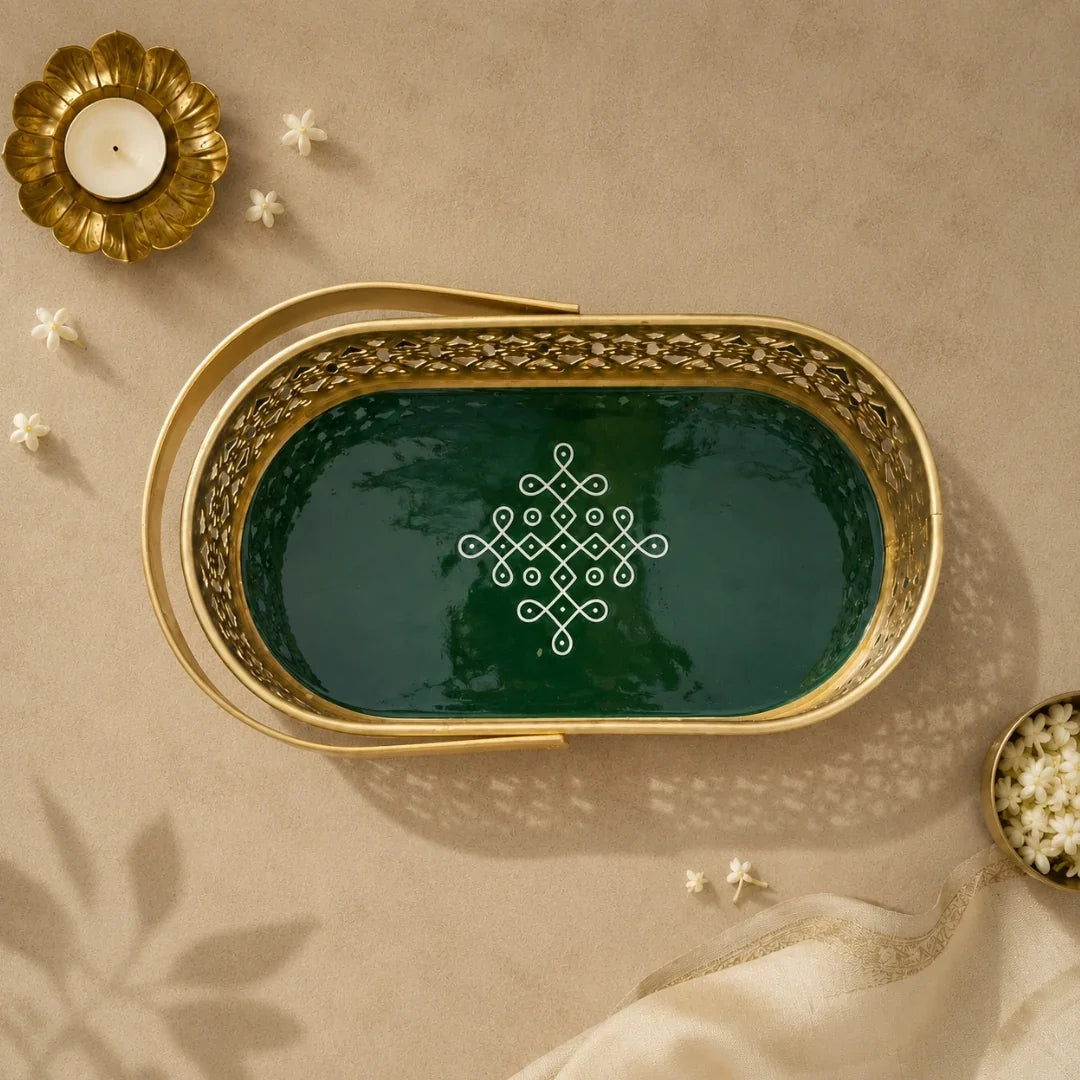 An oval decorative tray with a gold-toned metal frame and a dark green interior, featuring an intricate white geometric pattern in the center. The tray is styled in a flat-lay setting on a beige textured surface with a gold candle holder, small white flowers, and a cream fabric napkin.