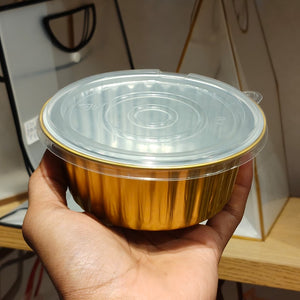 A hand holding a round disposable gold foil bowl with a clear plastic lid, shown in a real indoor setting with shelves and objects in the background.