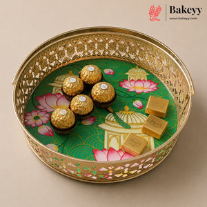 A round gold-toned decorative tray with intricate cutout detailing, holding five Ferrero Rocher chocolates and three gold-wrapped chocolate bars over a green printed insert with pink lotus motifs. The Bakeyy logo appears in the top right corner.