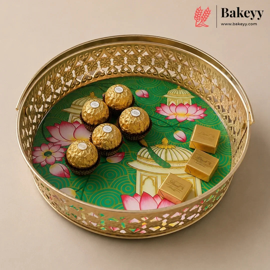 A round gold-toned decorative tray with intricate cutout detailing, holding five Ferrero Rocher chocolates and three gold-wrapped chocolate bars over a green printed insert with pink lotus motifs. The Bakeyy logo appears in the top right corner.