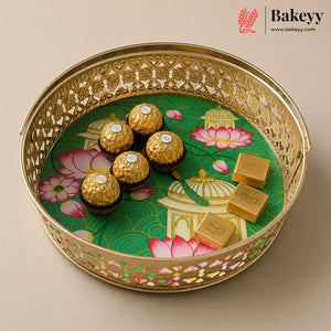 A round gold-toned decorative tray with intricate cutout detailing, holding five Ferrero Rocher chocolates and three gold-wrapped chocolate bars over a green printed insert with pink lotus motifs. The Bakeyy logo appears in the top right corner.