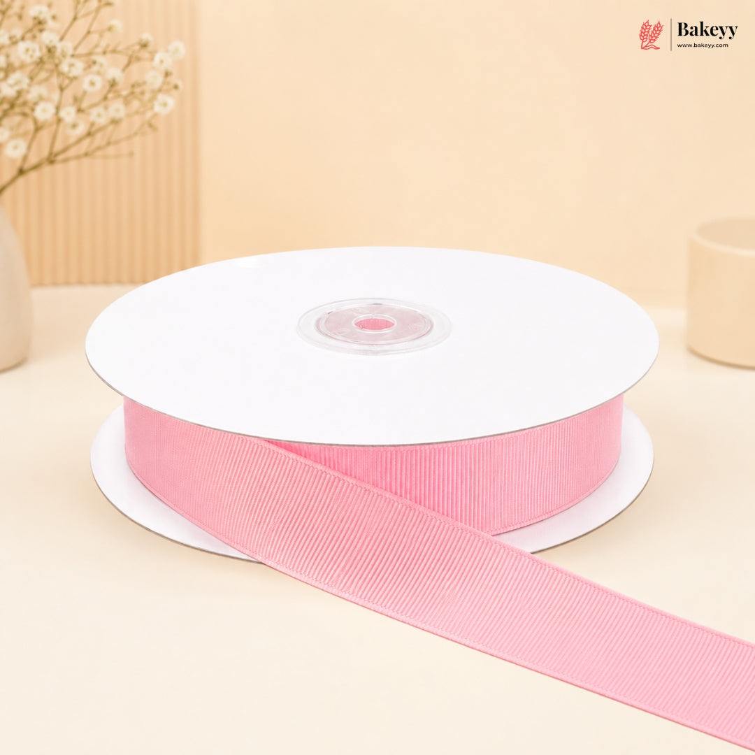 A spool of light pink grosgrain ribbon with a length draped out in front, set against a warm beige background with soft decorative props.
