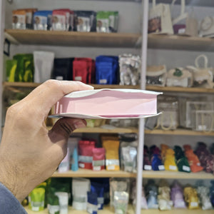 A hand holds a pink and white rectangular product, likely a book or notebook, in front of a blurred store shelf background filled with colorful packaged items.