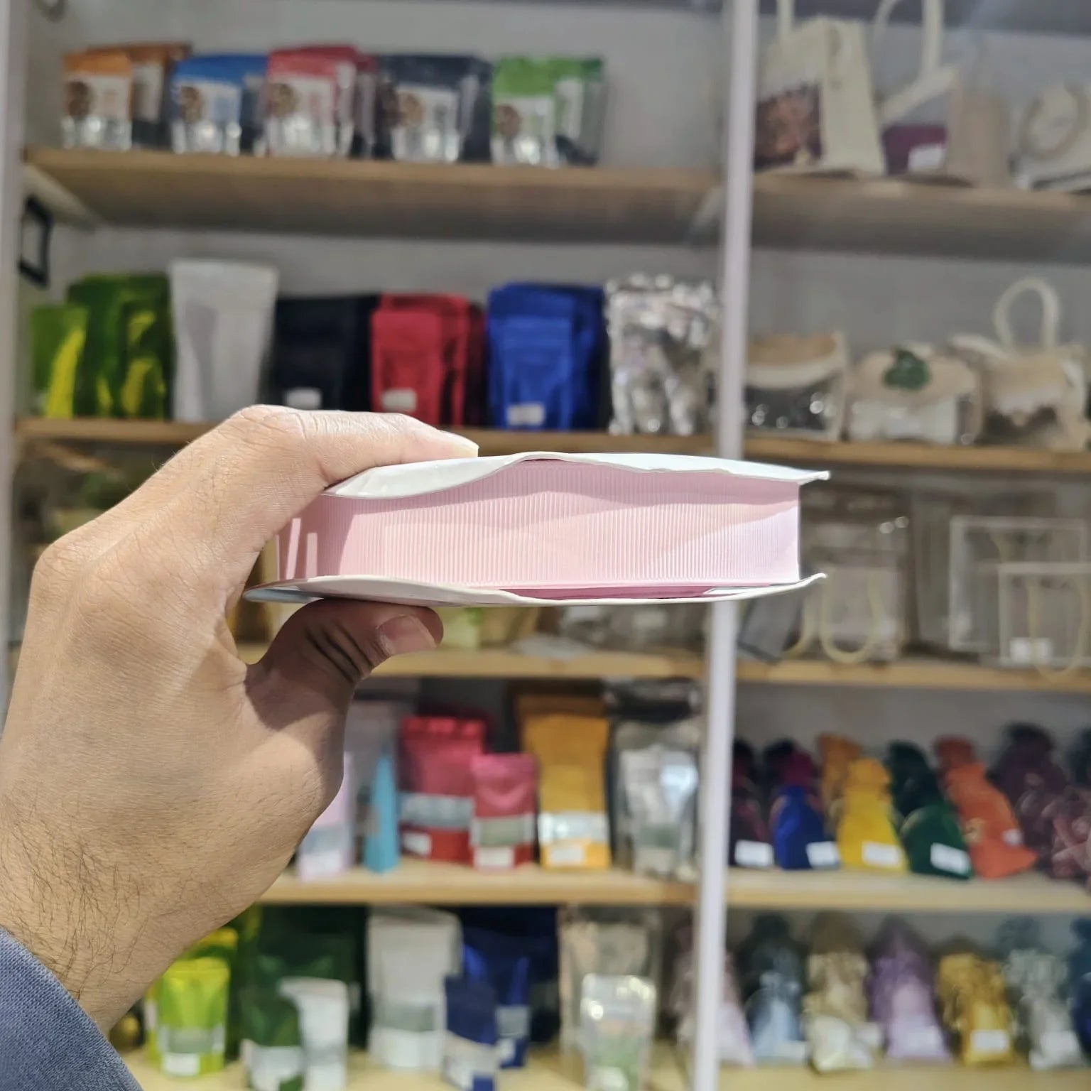 A hand holds a pink and white rectangular product, likely a book or notebook, in front of a blurred store shelf background filled with colorful packaged items.