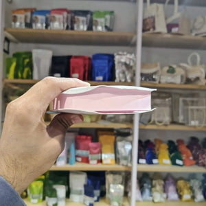 A hand holds a pink and white rectangular product, likely a book or notebook, in front of a blurred store shelf background filled with colorful packaged items.